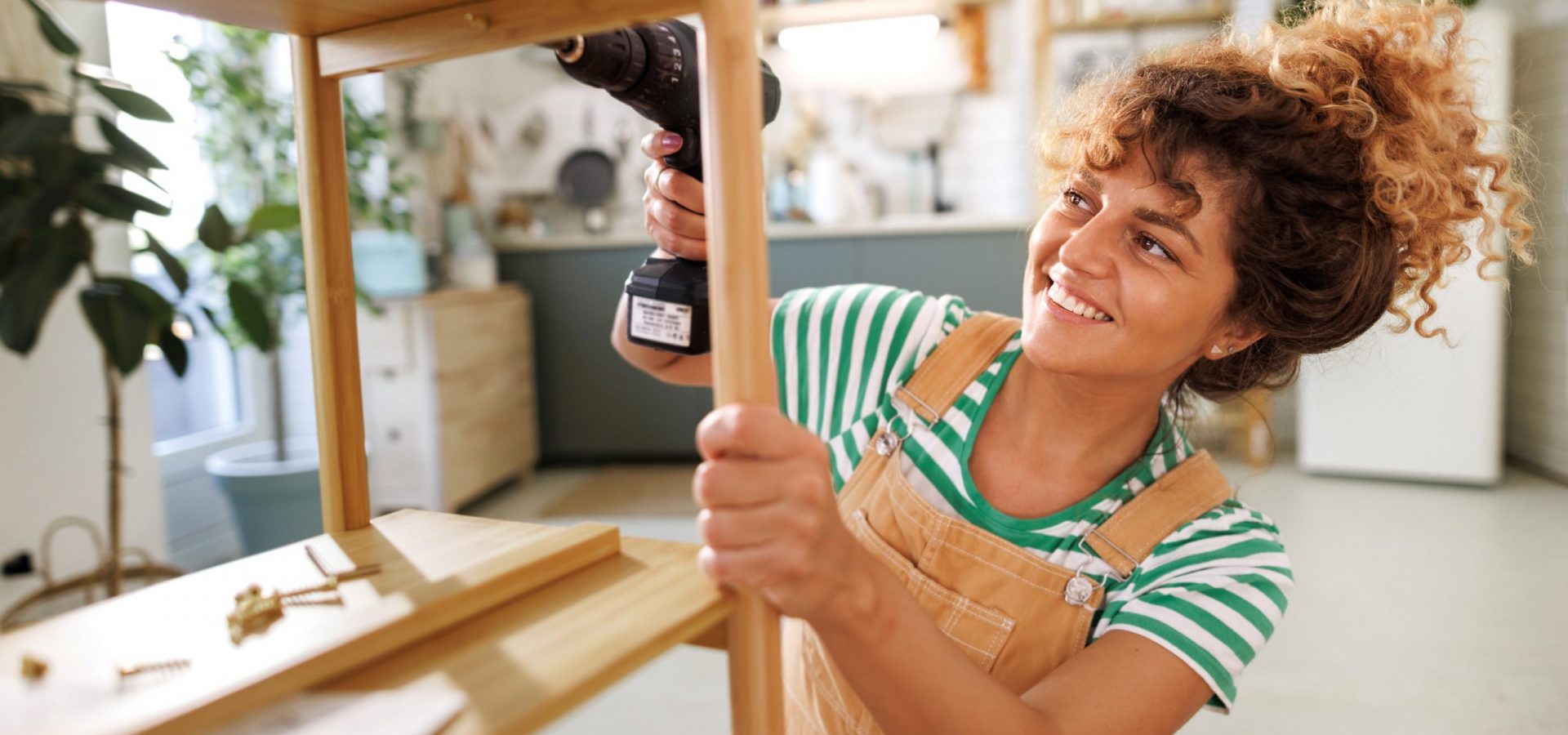 Young beautiful casually clothed woman assembling shelf with electric drill at home